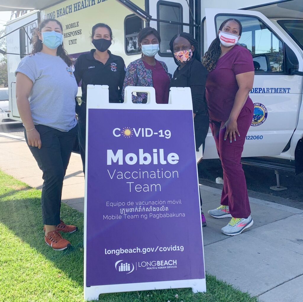 Five public health workers in masks stand in front of a mobile vaccination van. A large A-frame sign in front of them publicizes their services in English, Spanish, Khmer, and Tagalog.