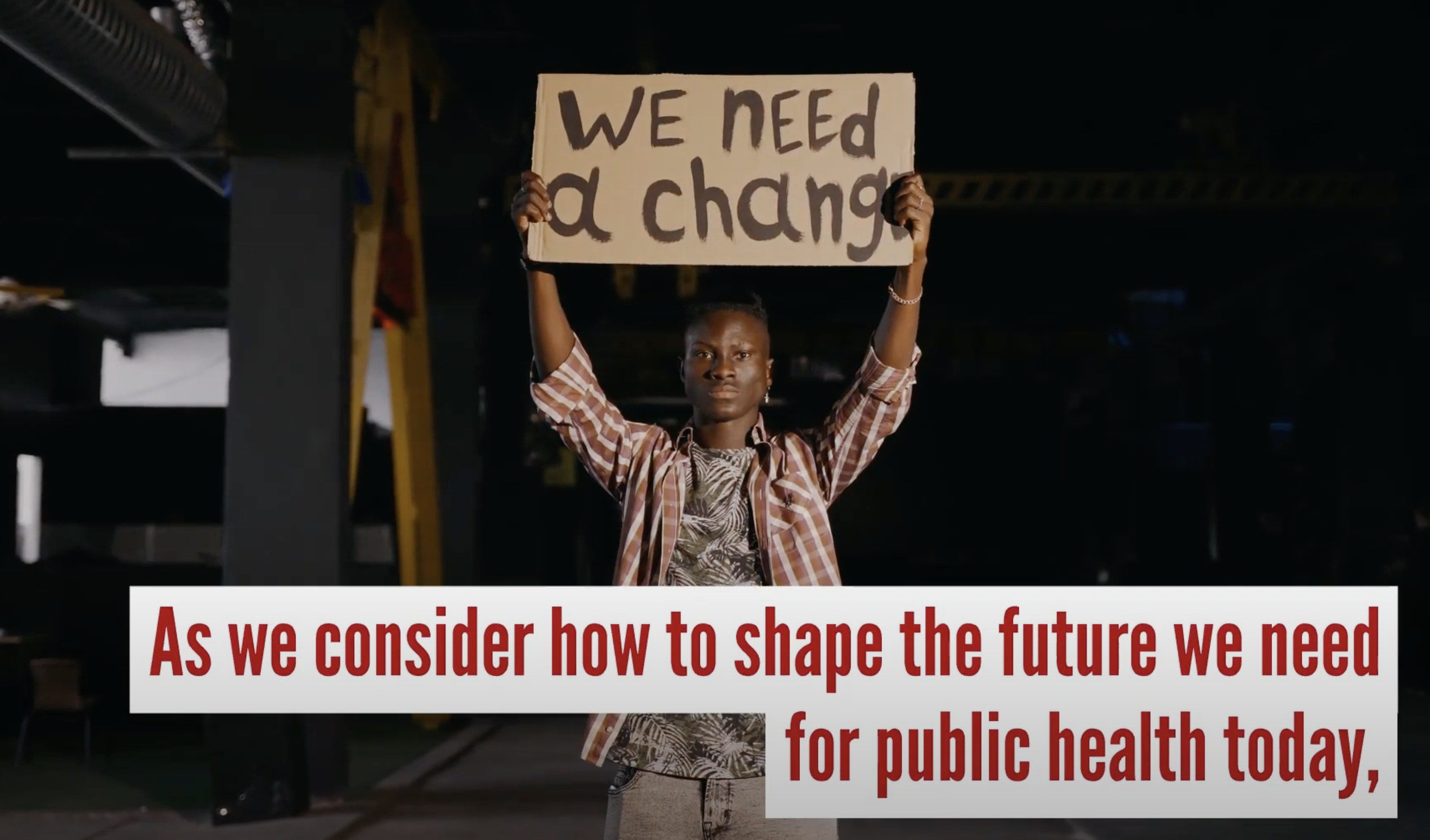Still from video shows young Black man holding placard above his head that reads "We need a change." Words superimposed on top say "As we consider how to shape the future we need for public health today,..."