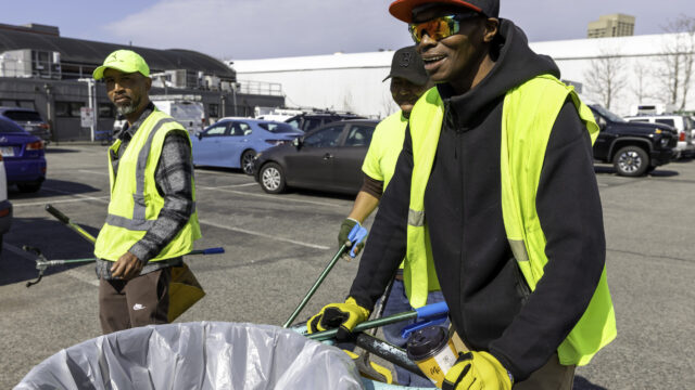 3 men in safety vests walking across a parking lot, one pushing a large trash receptacle