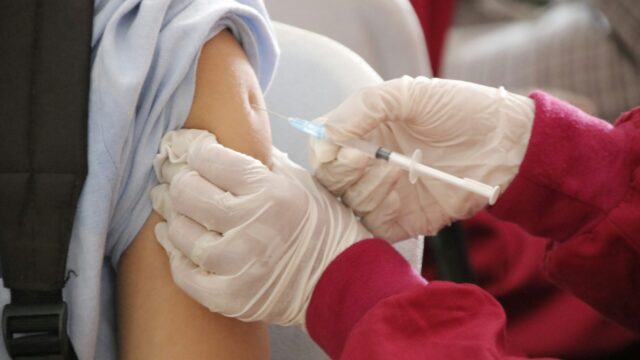 Image of a person wearing a black backpack receiving a vaccine from someone wearing latex gloves