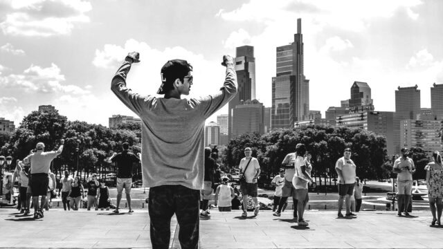 Young man with hands held over his head triumphantly, with people and city skyline in background. Photo by Prasad Panchakshari on Unsplash