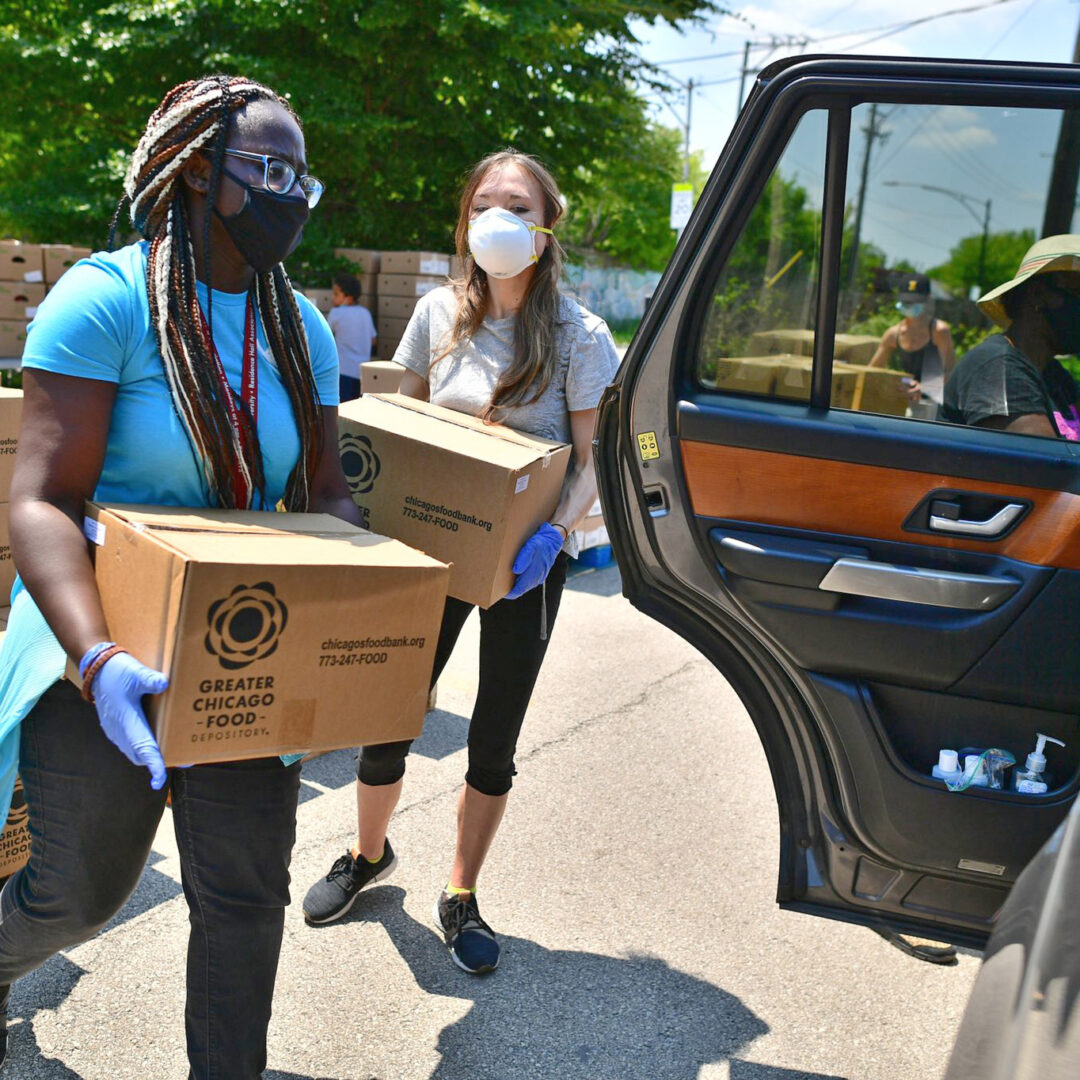 Two young women in masks carry boxes of food to load into a car