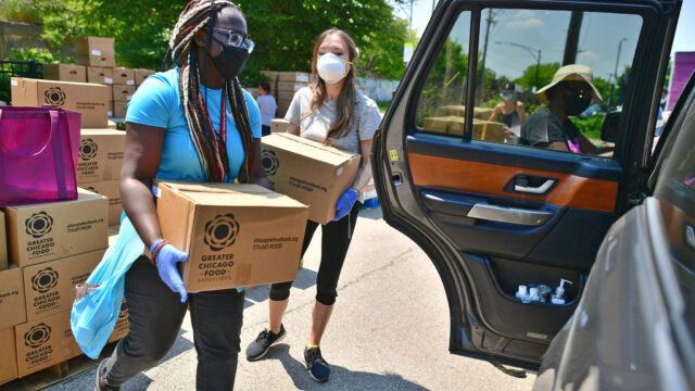Two young women in masks carry boxes of food to load into a car