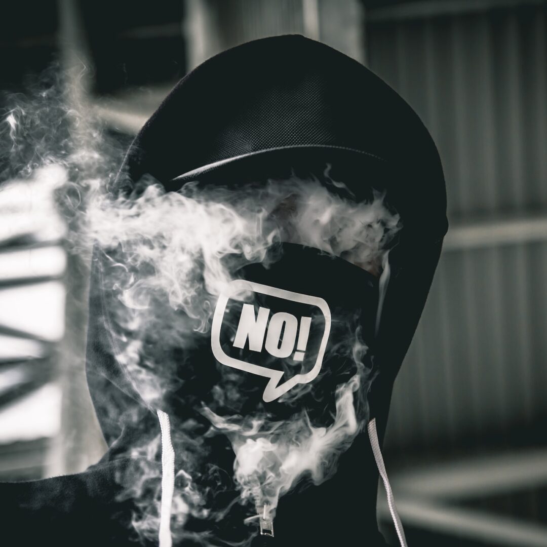 Hooded person vaping wearing mask that says NO. Photo by Matt Sirr on Unsplash.