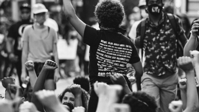 A woman of color with her back turned raises her fist in the air at a BLM protest. Photo by Gayatri Malhotra on Unsplash.