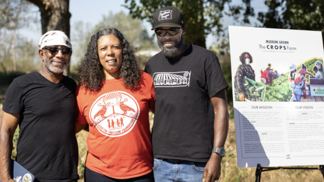 Three people from Mudbone Grown program standing outside next to a poster about the project.