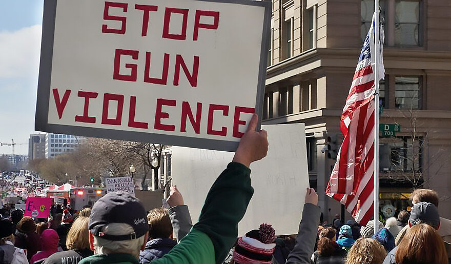 A person at a protest holding a sign stating, "STOP GUN VIOLENCE", next to an American flag. Photo by Chip Vincent.